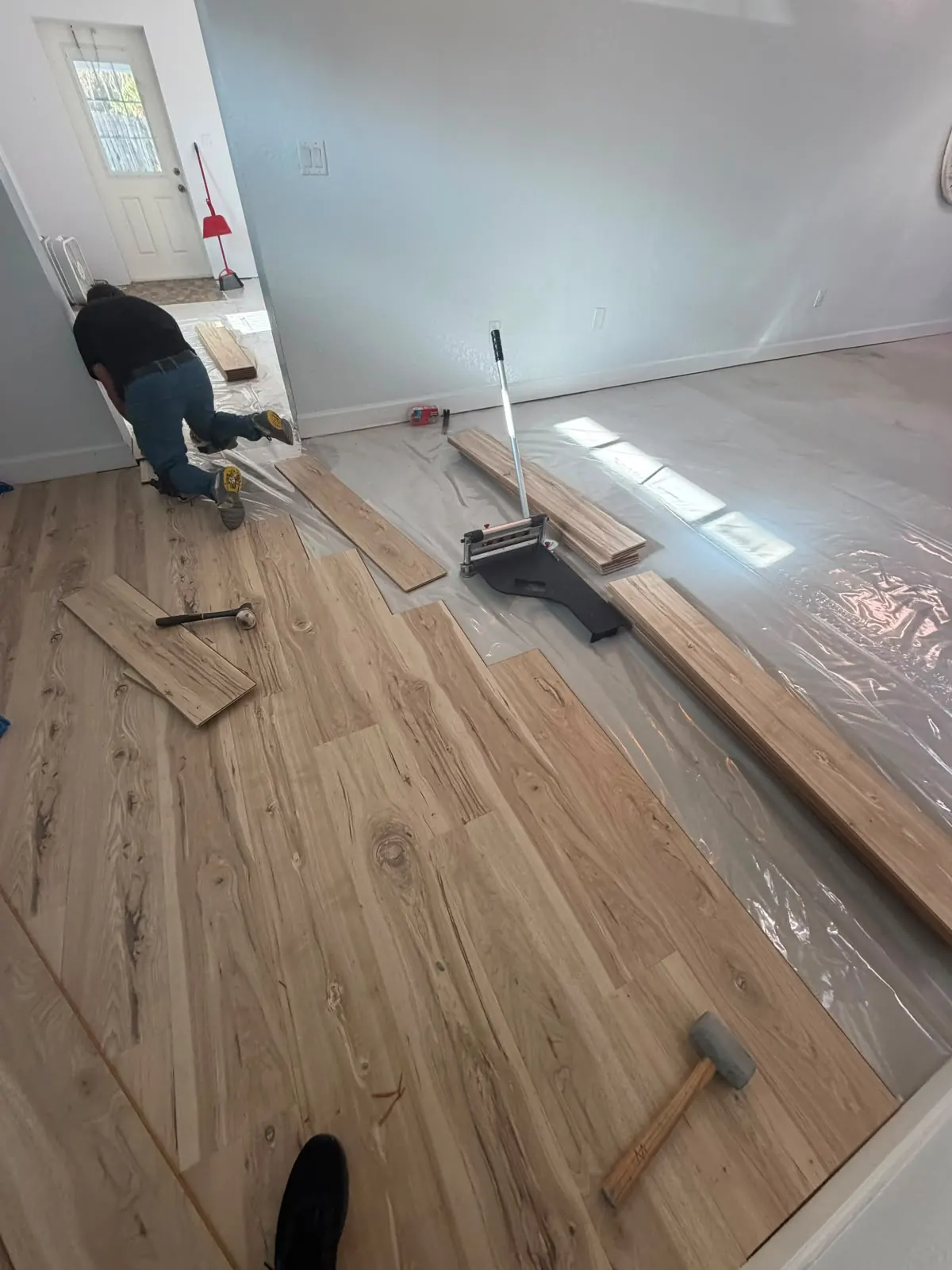 Installer kneeling while laying light wood-look plank flooring, with a plank cutter and loose planks on the floor