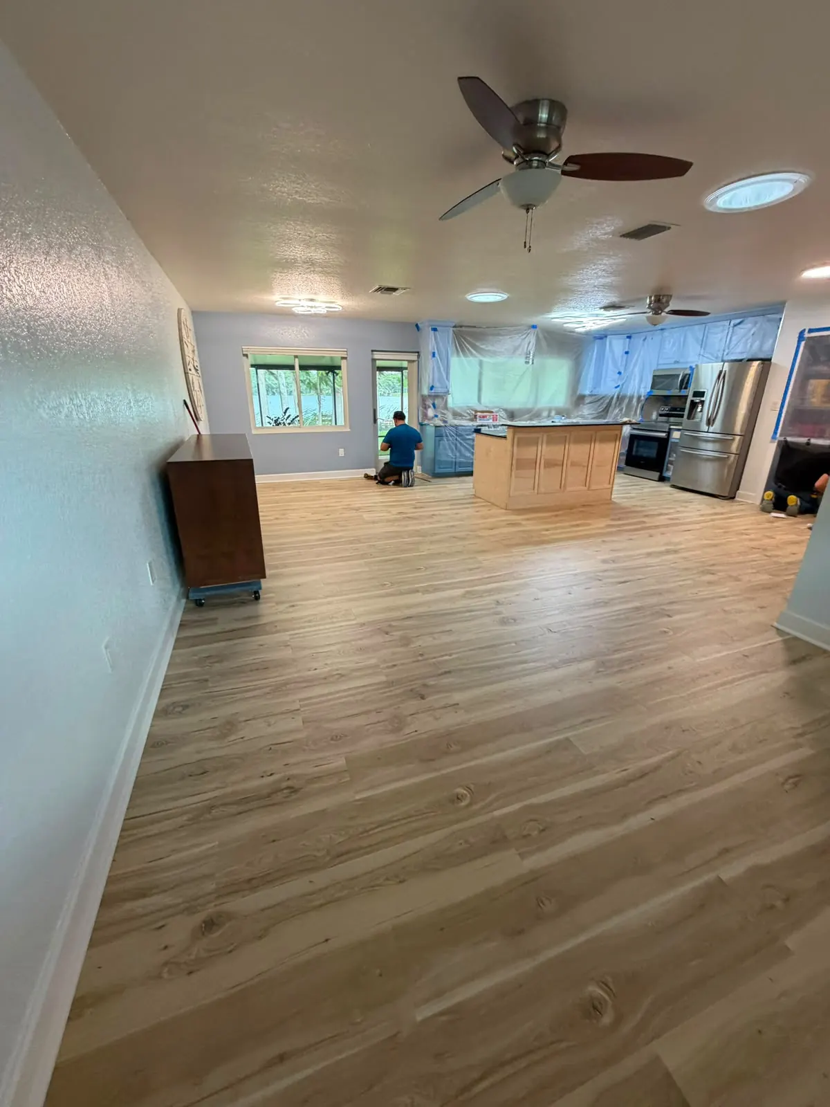 Light wood-look plank flooring across a living area that opens to the kitchen, with an installer working in the background